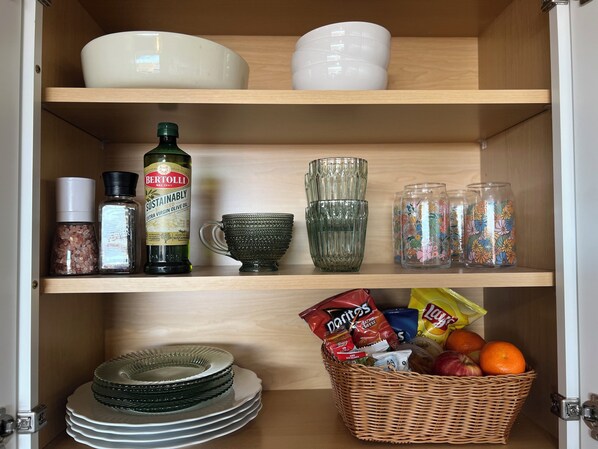 A kitchen cabinet with shelves containing various items including a bottle of extra virgin olive oil, salt and pepper shakers, a green glass mug, glass jars with floral designs, and a basket with snacks and fruit.