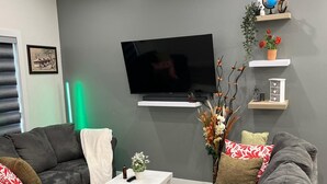 Ein modernes Wohnzimmer mit grauen Sofas, einem Flachbildfernseher, einem kleinen weißen Tisch und dekorativen Regalen mit Pflanzen und einem Globus.