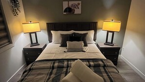 Ein ordentlich bezogenes Bett mit einer Karo-Decke und zwei Kissen, begleitet von zwei Nachttischen mit Lampen, in einem Zimmer mit dunkler Kopfteil und Wandbild.