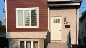 Ein zweistöckiges Haus mit brauner Fassade und einem beigen unteren Bereich. Eine weiße Tür mit kleinem Fenster darüber, sowie eine kleine Veranda mit Stufen, die zur Tür führen.