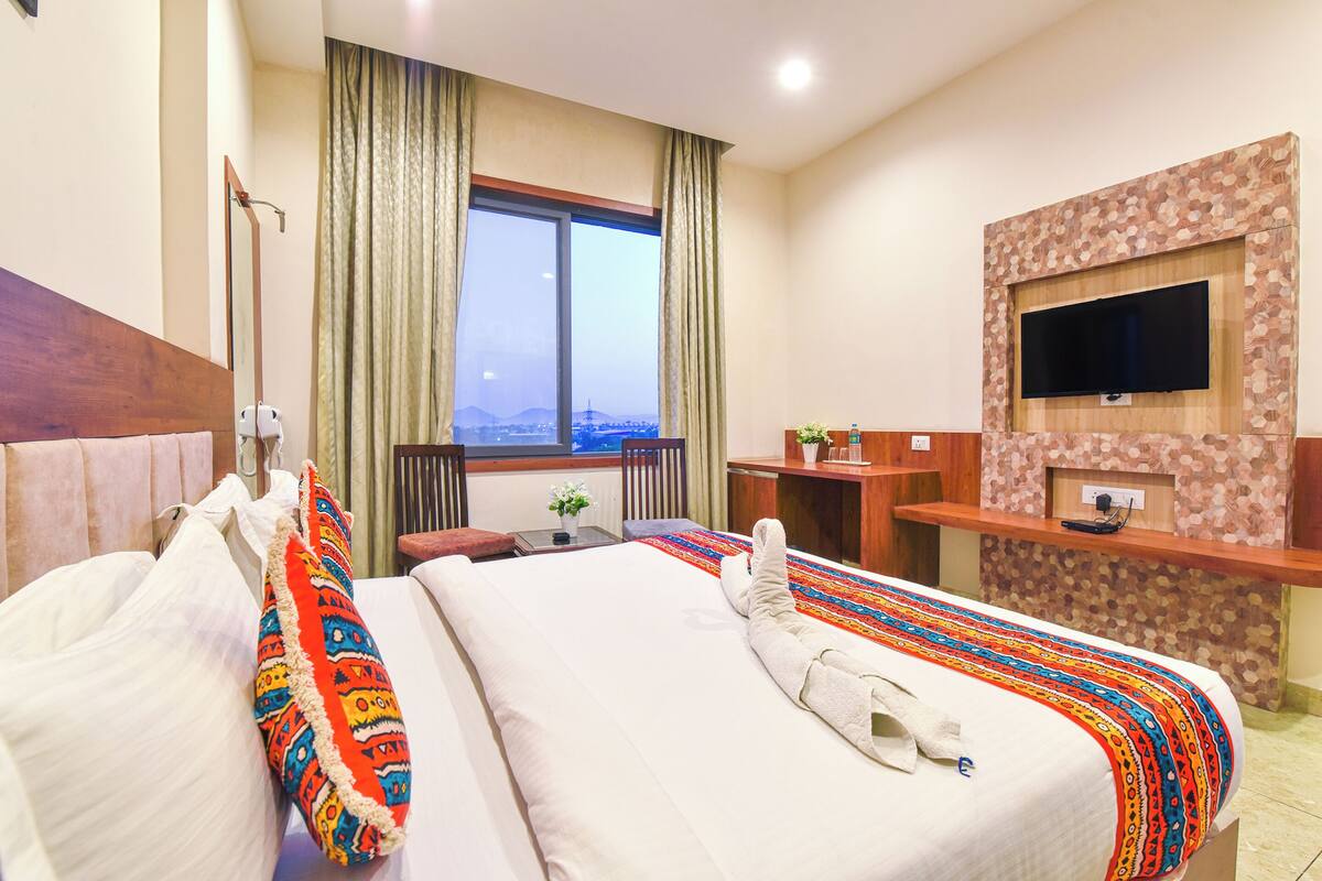 A hotel room with a large bed, a television mounted on a wooden wall, and a window with a view of a cityscape.