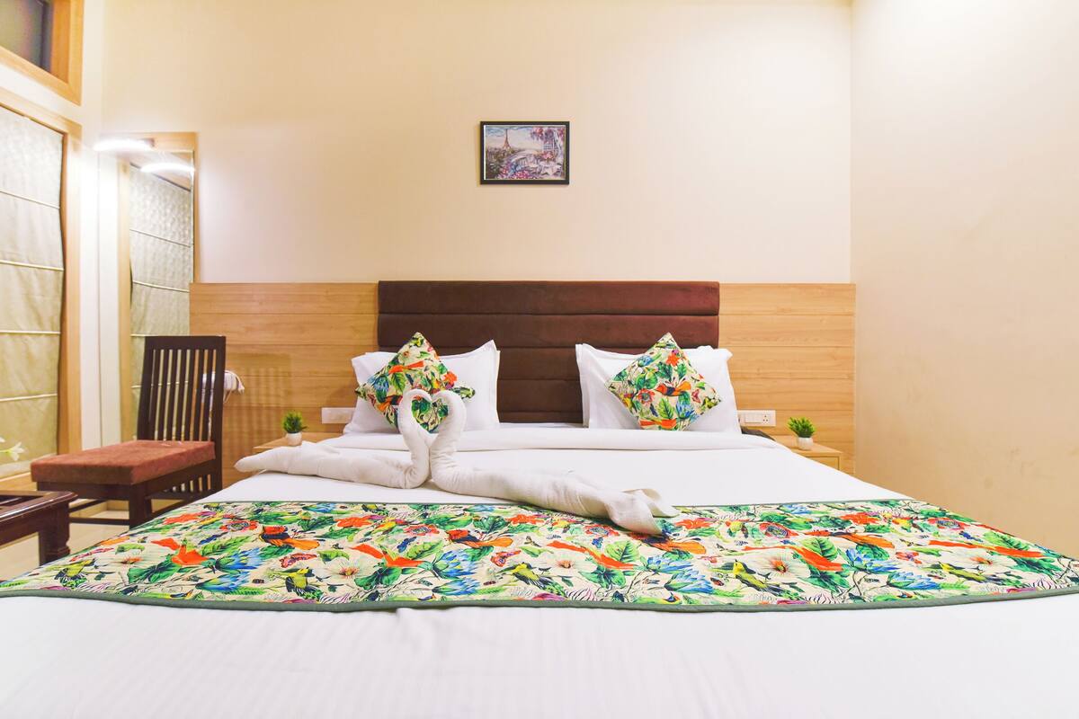 A neatly made bed with floral pillows and a colorful bedspread, a wooden headboard, a small bedside table with a plant, and a chair to the left.
