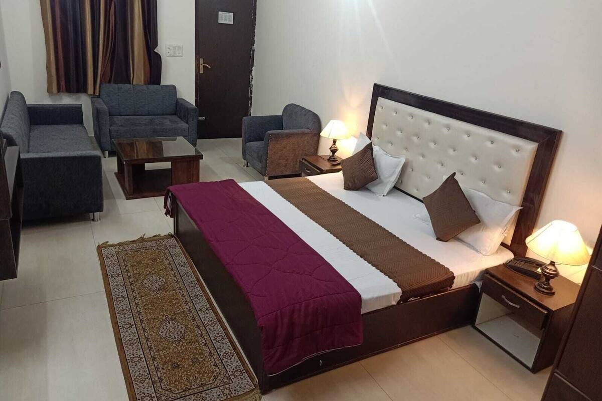 A hotel room with a bed, two armchairs, a sofa, a small table, a nightstand with a lamp, and a door.