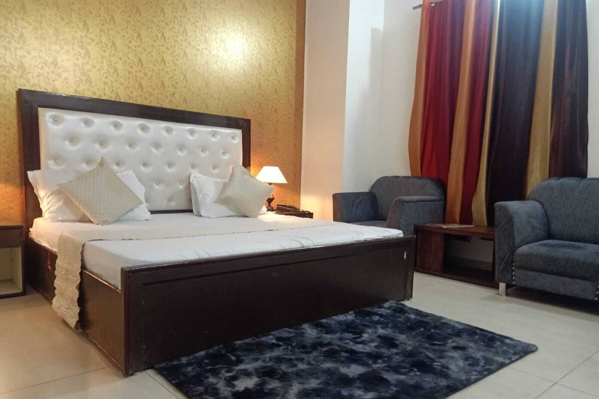 A bedroom with a large bed, a dark-colored headboard, a blue rug, a chair, and a nightstand with a lamp.