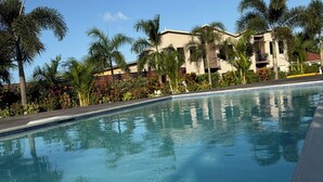 A clear swimming pool with blue-tiled edges, surrounded by palm trees and a building with balconies.