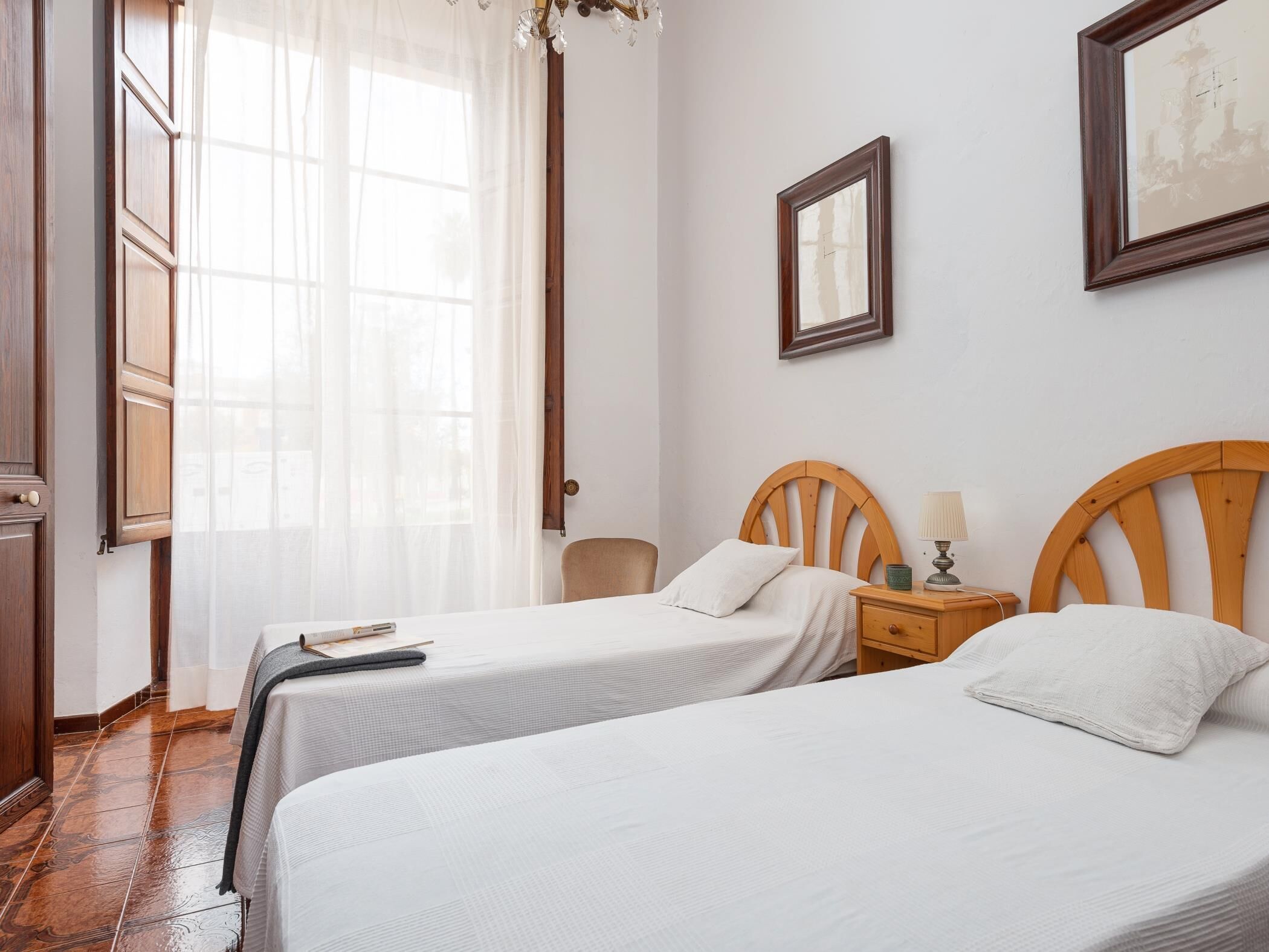 Habitación de hotel con dos camas individuales, una mesita de noche de madera con lámpara, un cuadro en la pared y una ventana con cortinas traslúcidas.