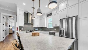 Una cocina moderna con armarios blancos, una isla grande, electrodomésticos de acero inoxidable e iluminación colgante.
