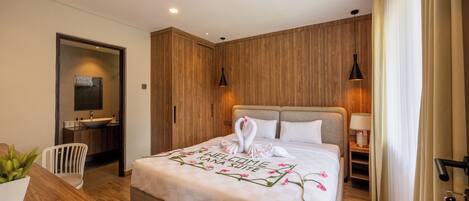 A modern hotel room with a wooden wall, a bed with a welcome mat, a bedside table, a chair, a desk, and a bathroom with a sink and mirror.