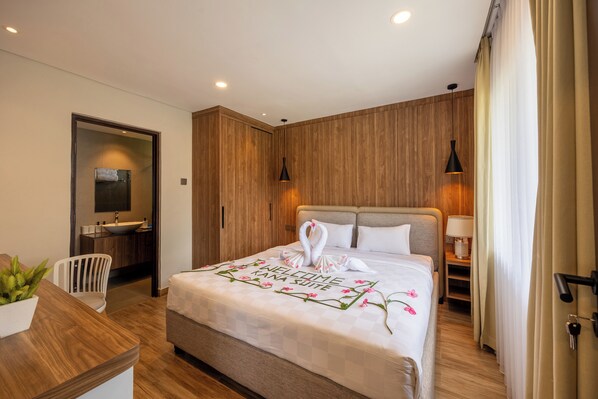 A modern hotel room with a wooden wall, a bed with a welcome mat, a bedside table, a chair, a desk, and a bathroom with a sink and mirror. - Kana Suites Nusa Dua By Fays Hospitality (Nusa Dua)