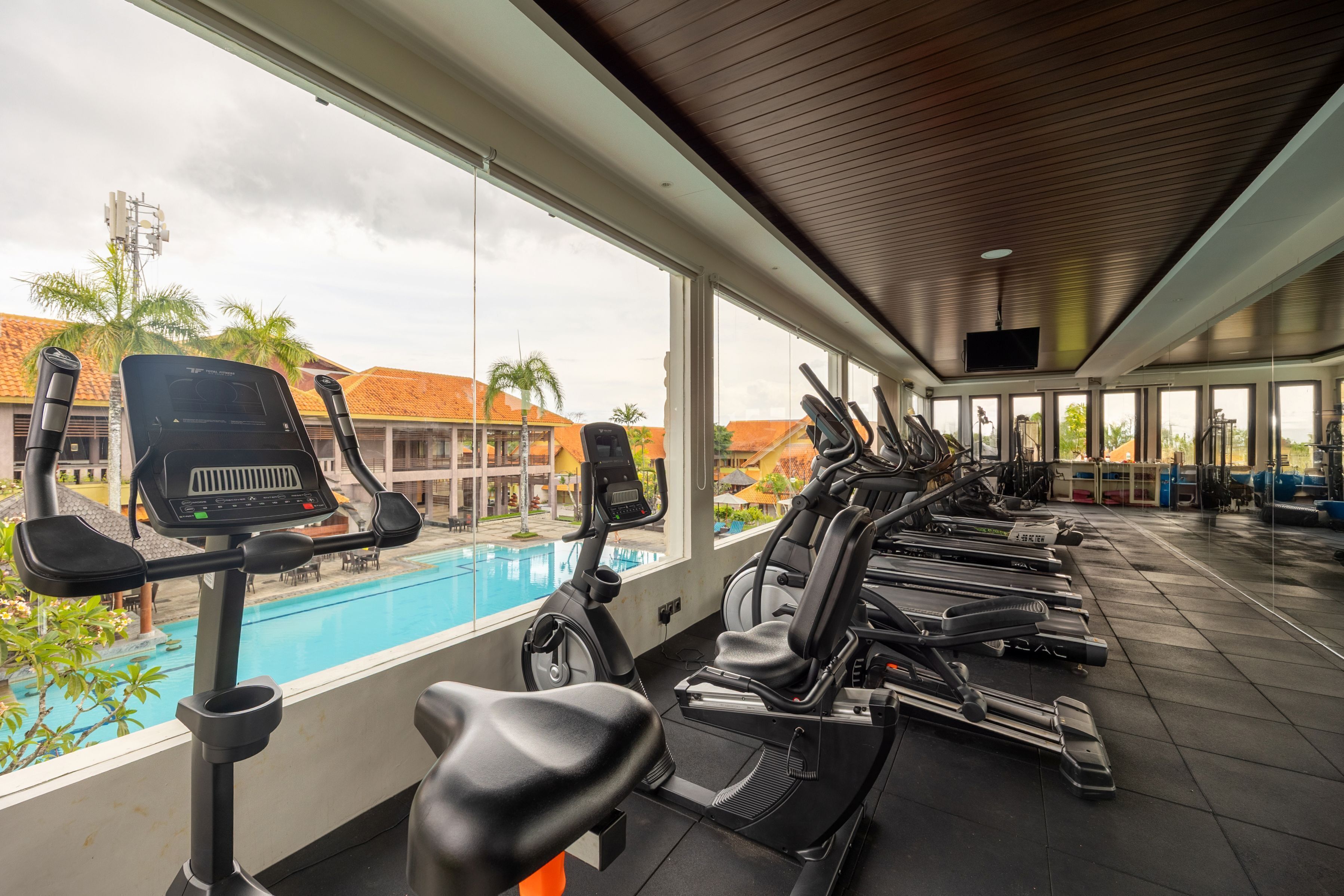 A gym with multiple treadmills and exercise bikes, large windows overlooking a swimming pool and palm trees.