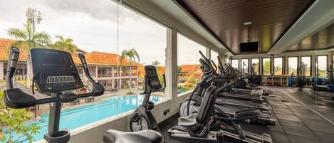 A gym with multiple treadmills and exercise bikes, large windows overlooking a swimming pool and palm trees.