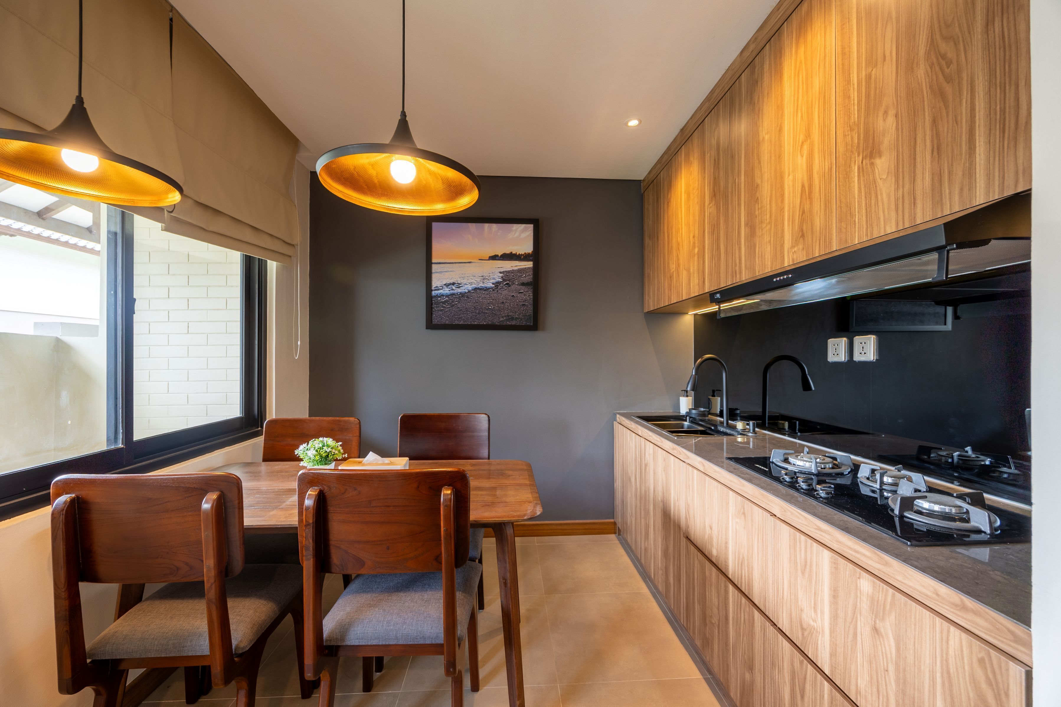 Una cocina moderna con armarios de madera, un comedor con mesa y sillas, lámparas colgantes y vistas a la playa a través del ventanal.