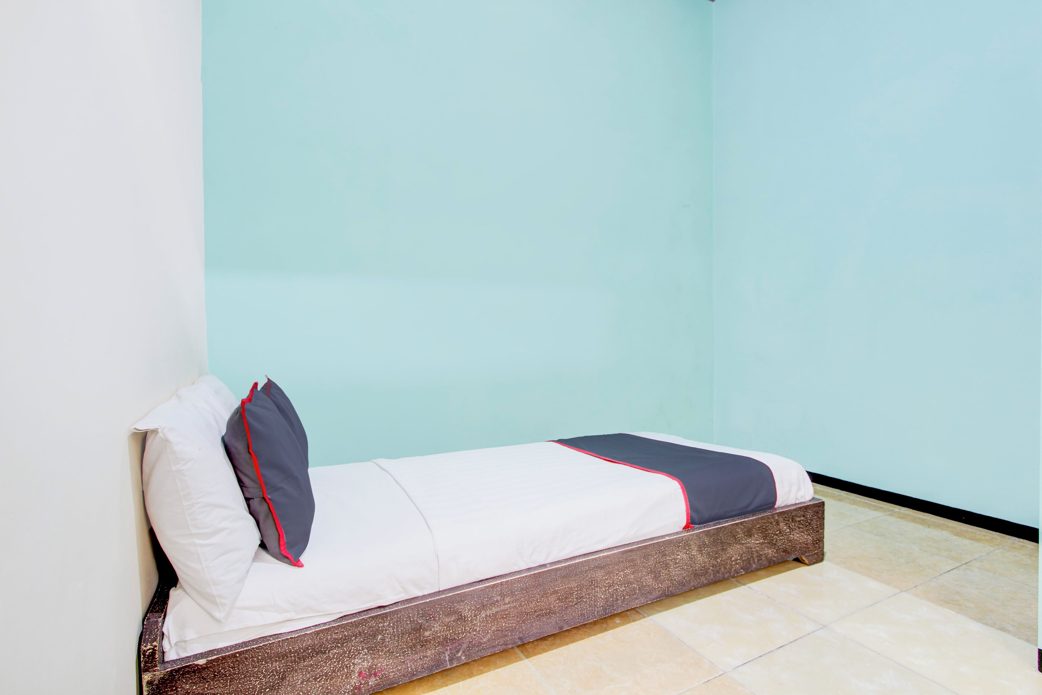 A single bed with a wooden frame, white bedding with red trim, and a light blue wall in the background.