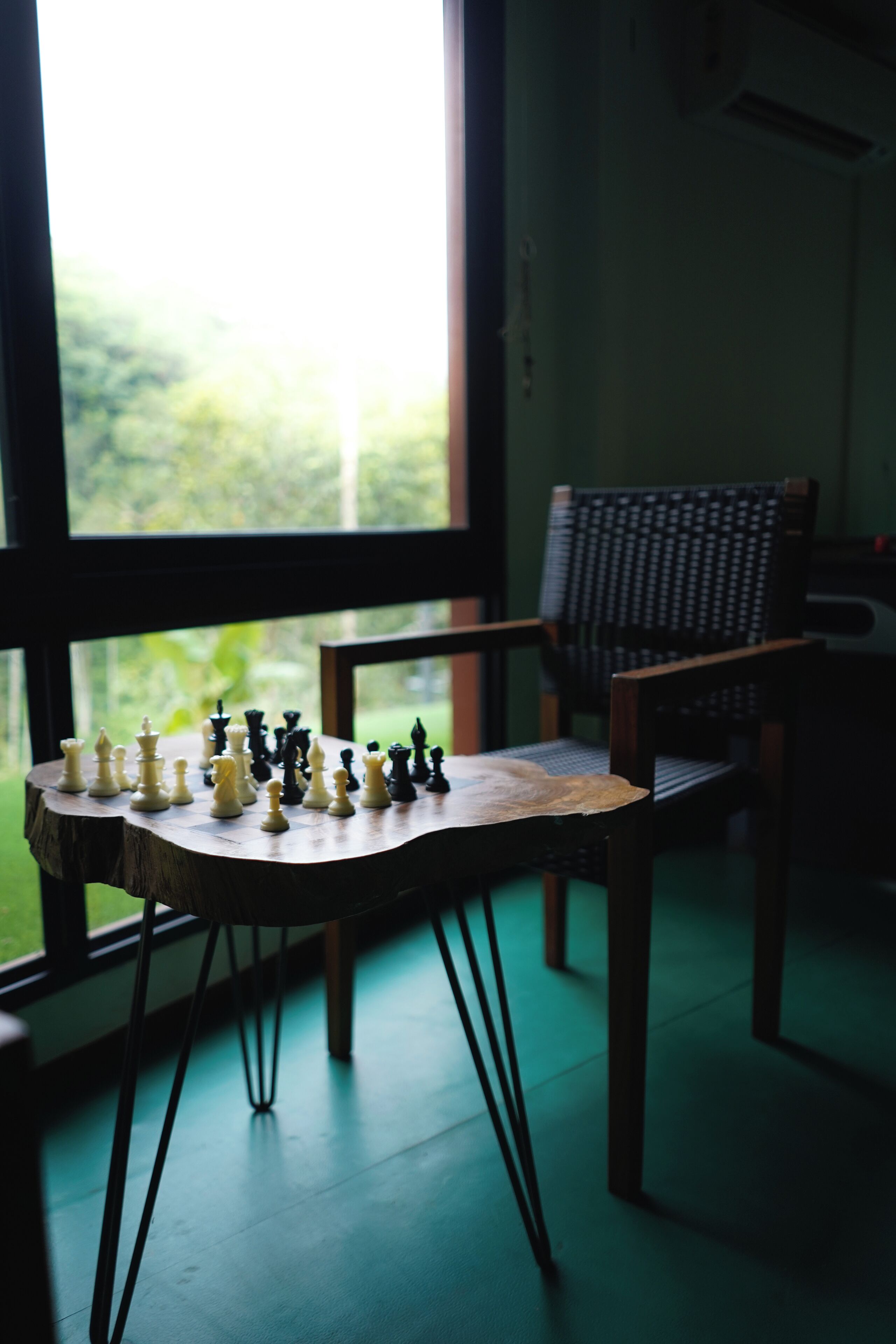 A chessboard on a table by a window with a chair nearby.