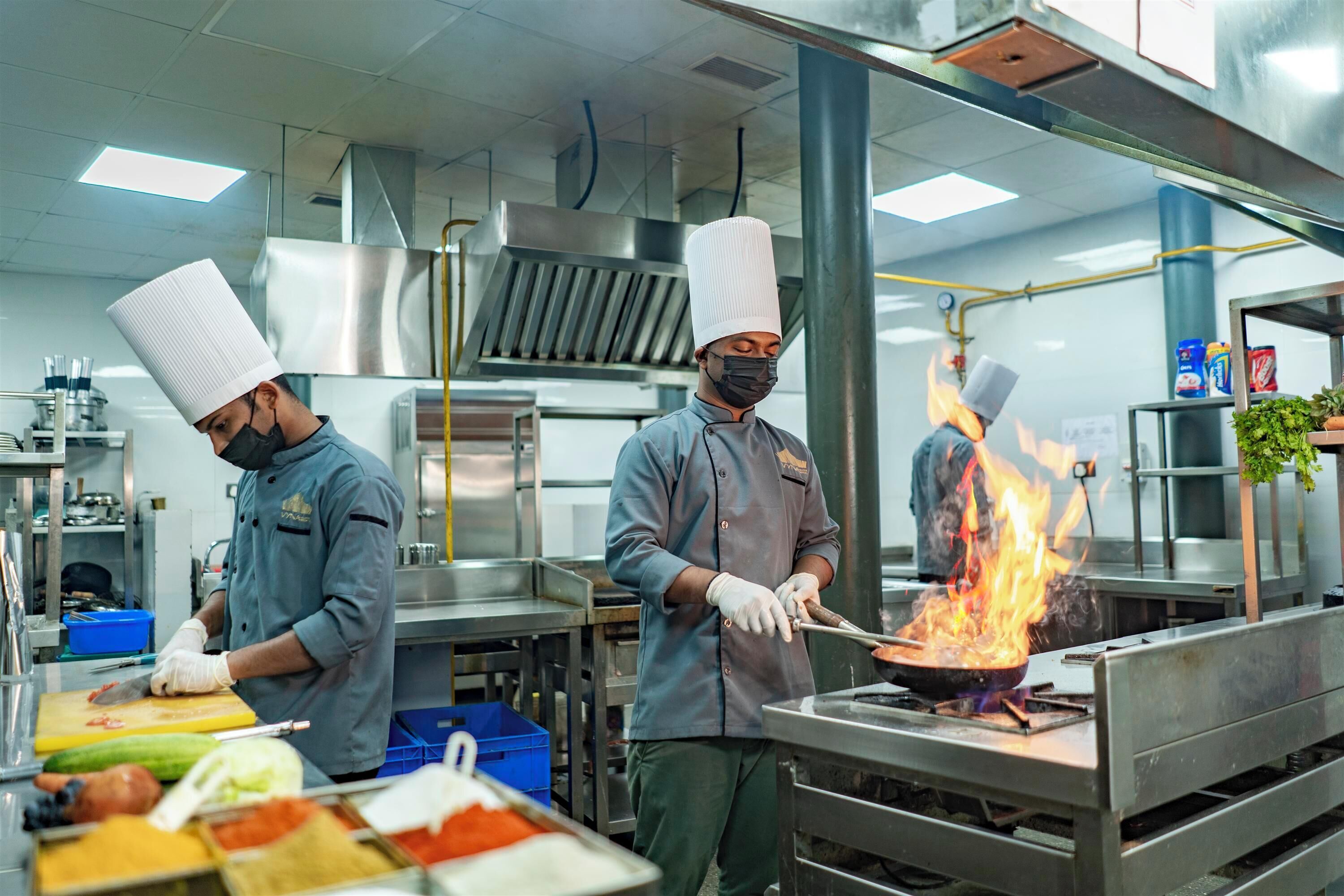 Une cuisine professionnelle où des chefs préparent des plats, avec des équipements en acier inoxydable et une variété d’ingrédients disposés sur le plan de travail.