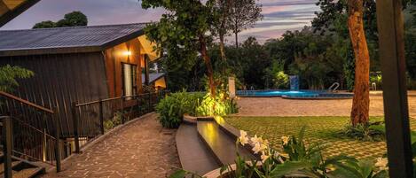 A stone pathway leading to a building with a metal railing, surrounded by lush greenery and a pool area.