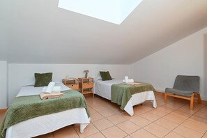 A bedroom with two beds, each with a green blanket and pillows, a wooden nightstand with a vase, and a gray chair.