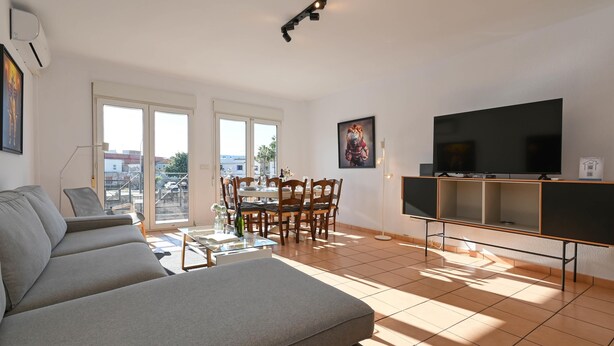 Un soggiorno con divano, tavolo da pranzo con sedie, televisione e balcone con vista su edifici e palme.