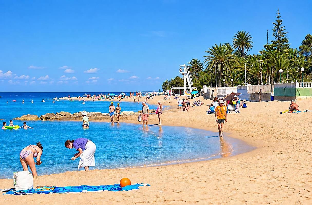 Una platja amb gent nedant i passejant, palmeres i una torre de socorristes.