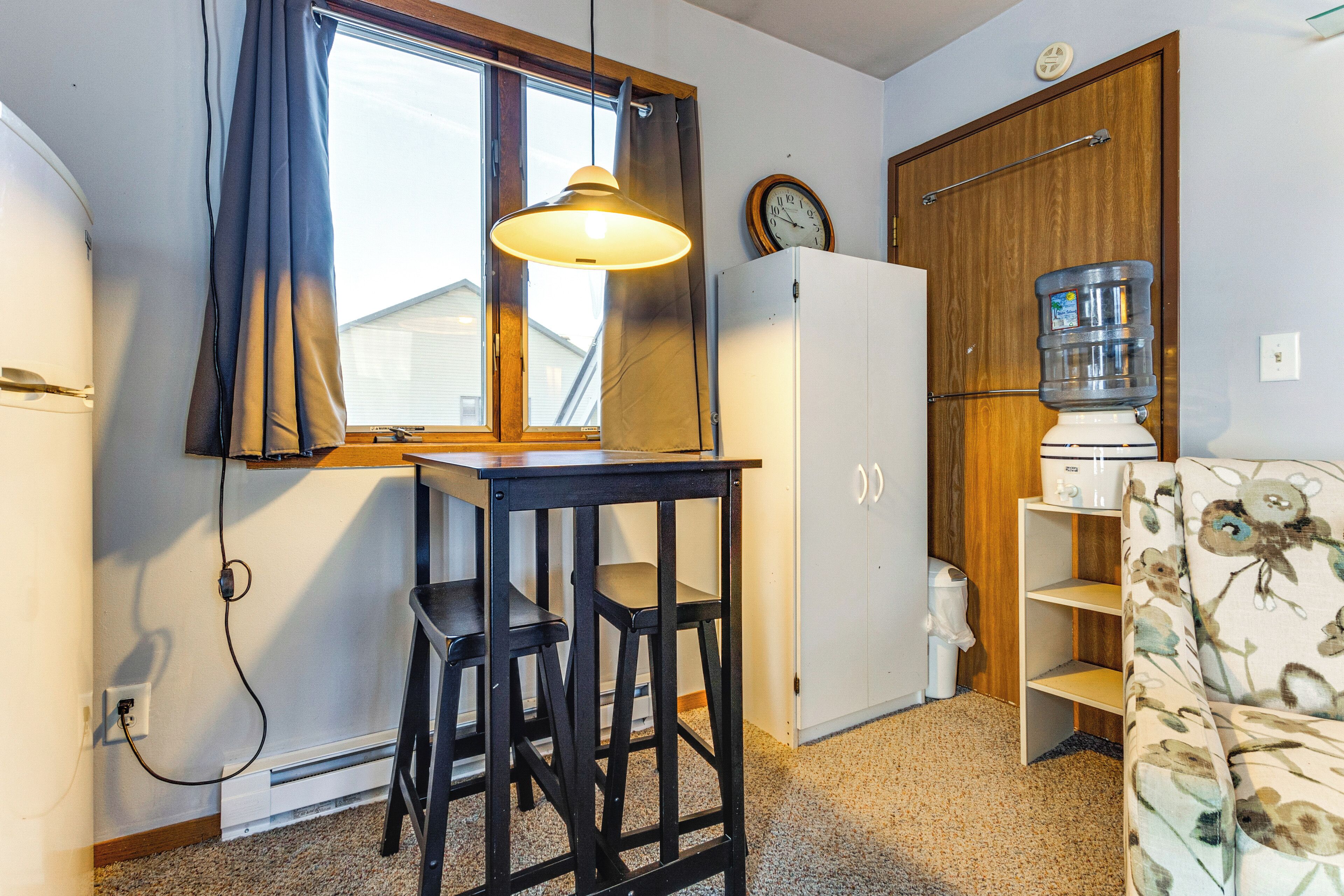 A compact kitchen with a window, a refrigerator, a water dispenser, a clock, and a floral sofa.