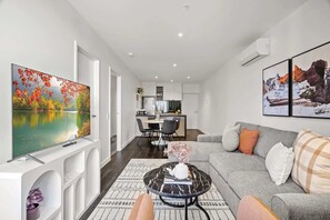 A modern living room with a flat-screen TV, a sofa, a coffee table, and a dining area in the background.