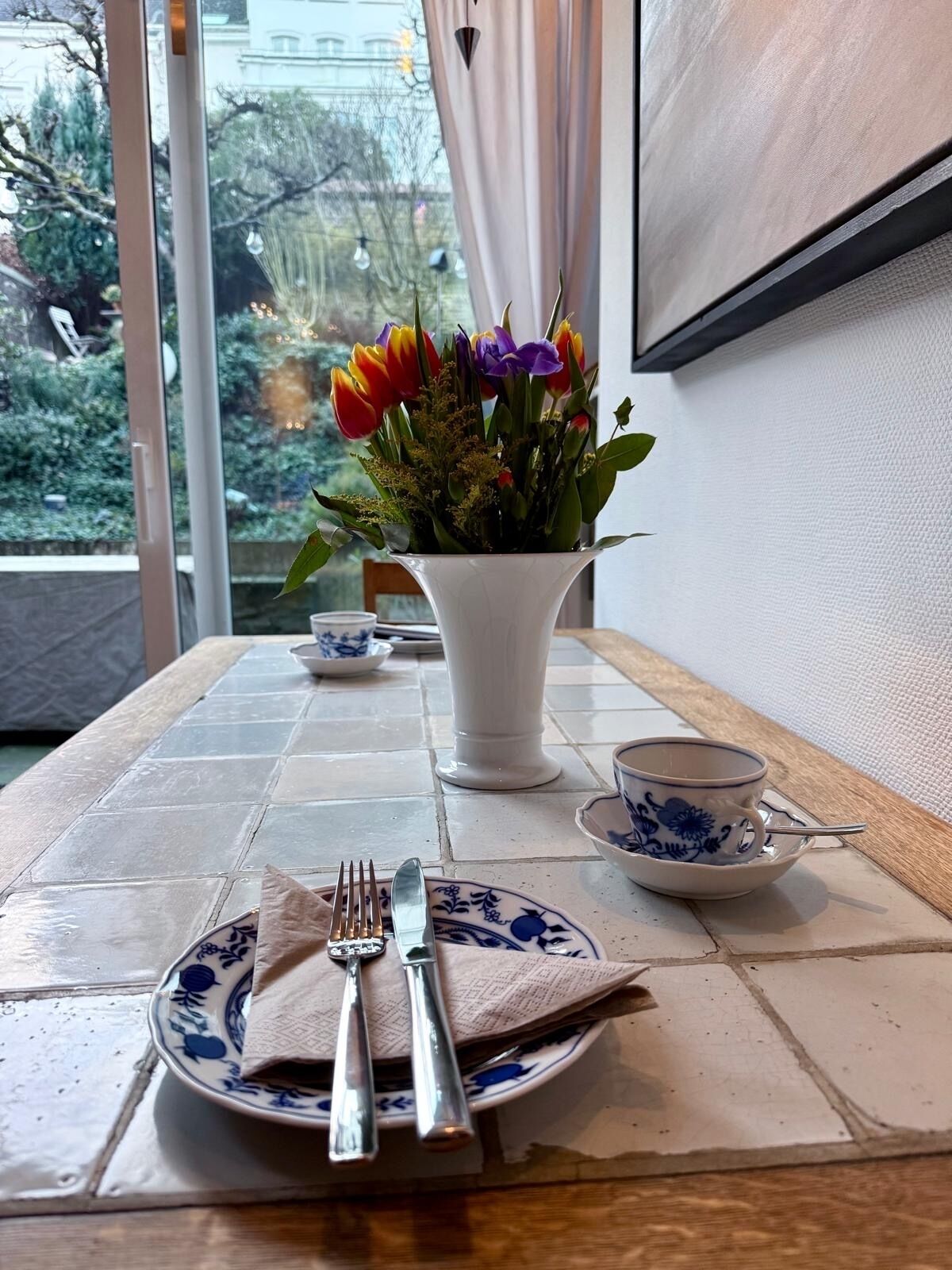 Una mesa de comedor con un jarrón blanco de tulipanes coloridos, un plato con cubiertos y una taza azul y blanca sobre un suelo de baldosas.