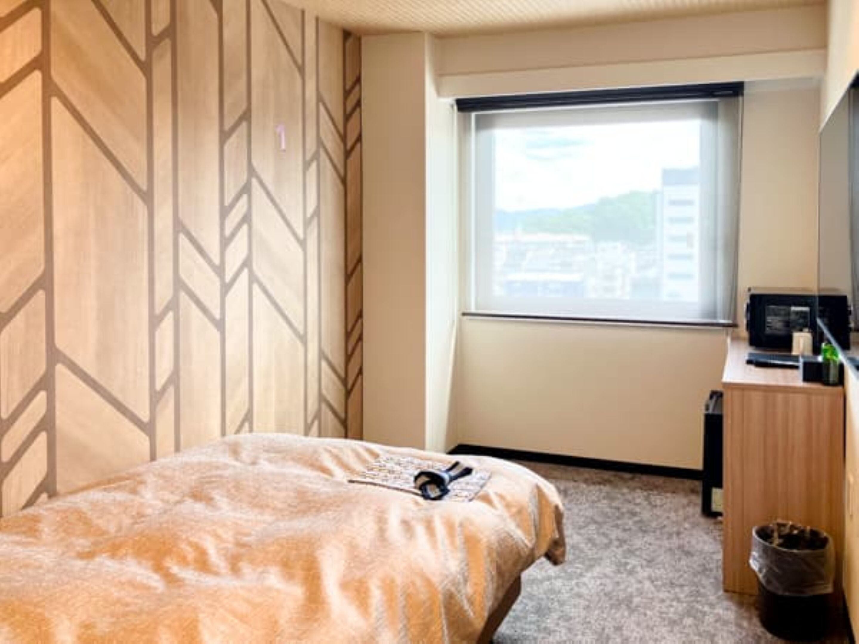 A hotel room with a bed, a desk with a computer, a trash bin, and a window with a view of buildings.