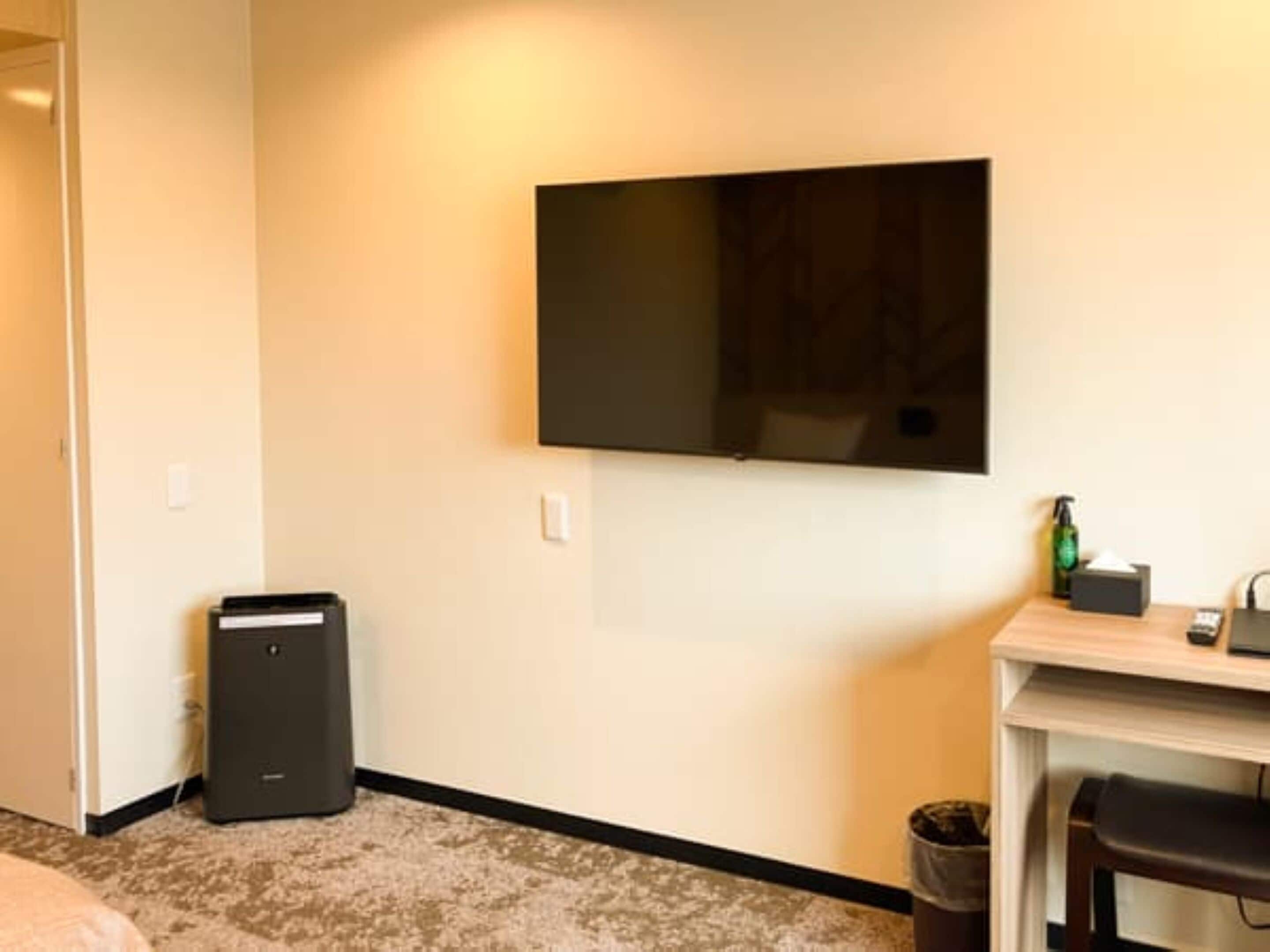 Ein Hotelzimmer mit flachbildfernseher an der Wand, einem kleinen Tisch mit Flasche und Taschentuchbox, einem Mülleimer und einem Stuhl.