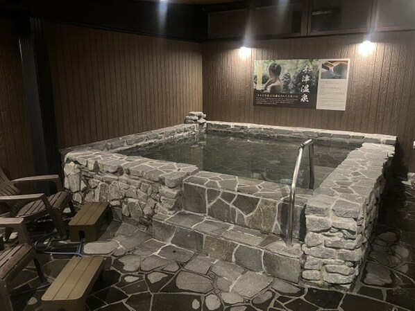 A stone hot tub with a metal ladder, surrounded by wooden walls and stone flooring, with chairs and a wooden bench nearby.