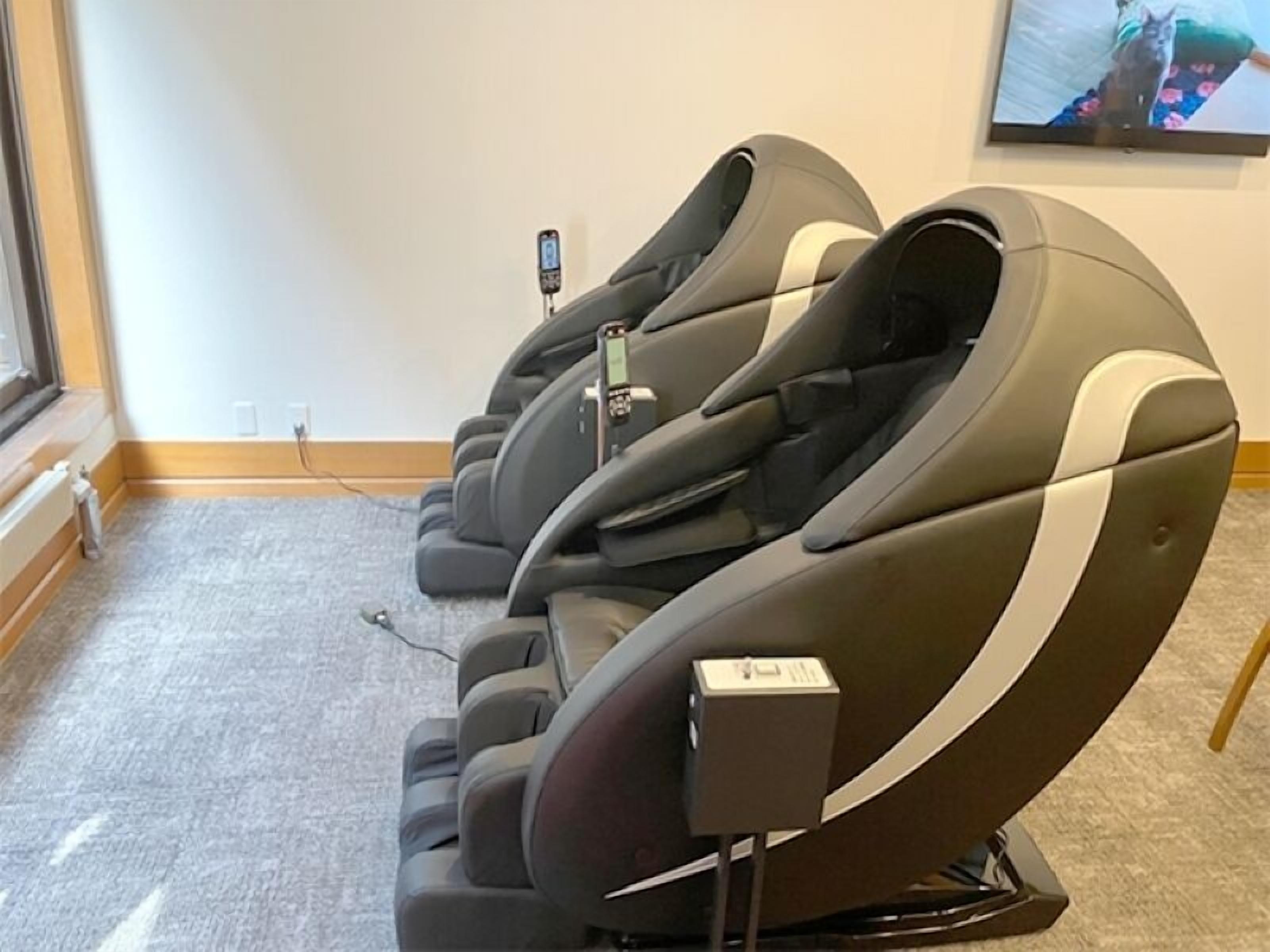 Reihenweise Massage-Stühle in einem Raum mit Teppichboden und Fernseher an der Wand.