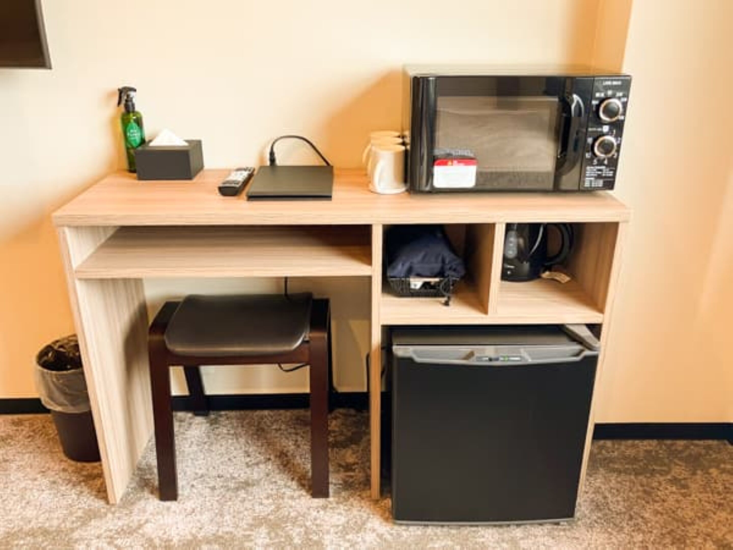 A compact hotel room with a wooden desk, a black refrigerator, a microwave, a chair, and a trash bin.