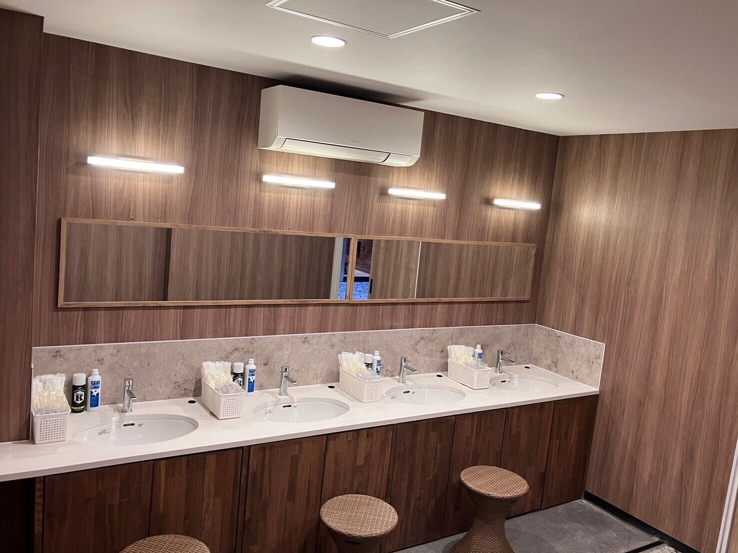 A modern bathroom with wooden paneling, a large mirror, two sinks, and three wicker stools.