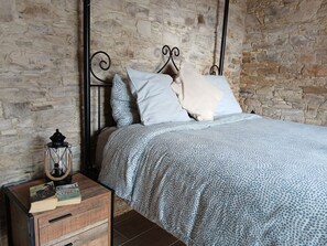 Un dormitorio con pared de piedra, cama con ropa de cama estampada, mesita de noche con libros y un farol.