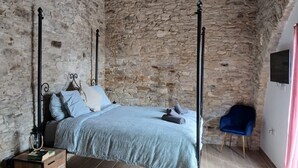 Une chambre avec un mur en pierre, un lit à baldaquin, une table de chevet en bois, une chaise bleue et un téléviseur fixé au mur.