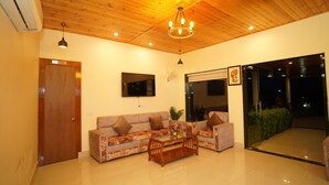 A living room with a wooden ceiling, a sofa set, a glass coffee table, and a TV mounted on the wall. - Vasant Corbett Resort (Ramnagar)