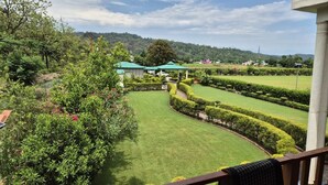 A landscaped garden with neatly trimmed hedges and a lawn, a building with a green roof, and a view of a hilly landscape. - Vasant Corbett Resort (Ramnagar)
