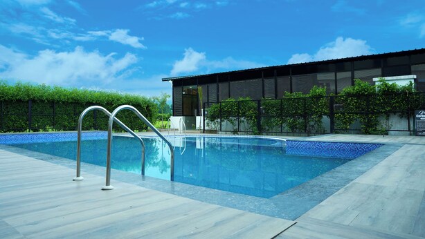 A swimming pool with metal ladders, surrounded by a tiled deck and a modern building with large windows. - Vasant Corbett Resort (Ramnagar)
