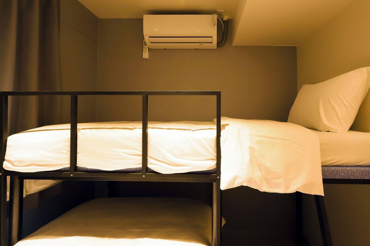 Ein Etagenbett mit Kopfstütze, Kissen und Decke. Eine Klimaanlage ist an der Wand darüber angebracht.