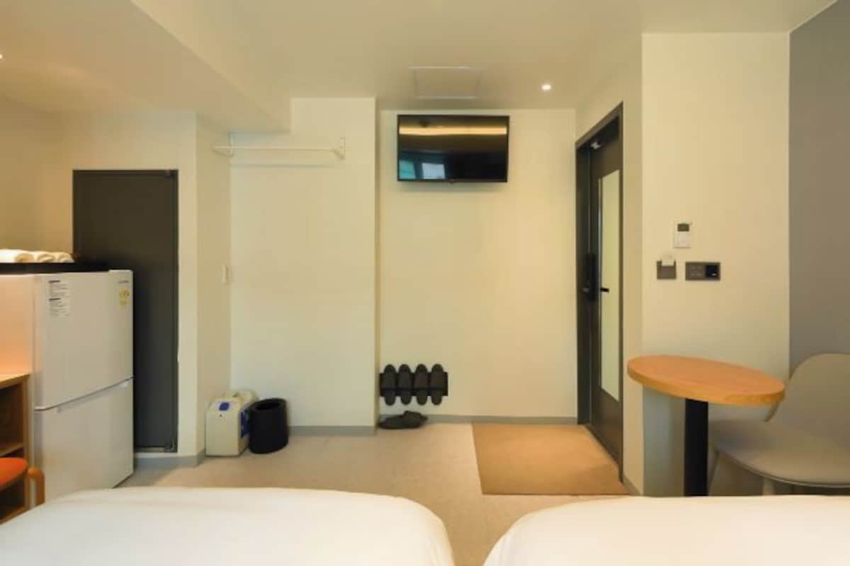 Habitación de hotel con dos camas, una mesita, televisor montado en la pared, refrigerador y un zapatero.