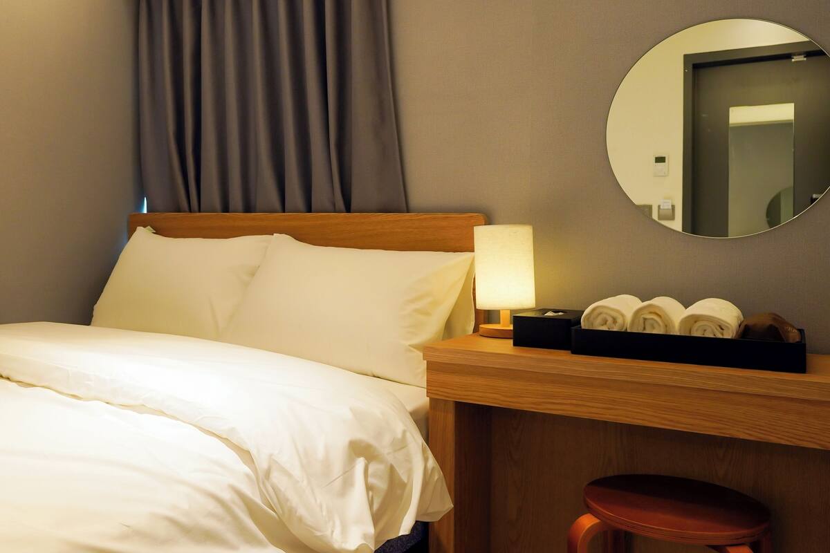 Ein Hotelzimmer mit einem ordentlich bezogenen Bett, einem Nachttisch mit Lampe, einem Spiegel und einem kleinen Hocker.