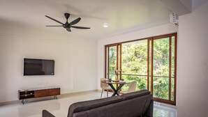 Una sala de estar moderna con un sofá, un televisor montado en la pared, un ventilador de techo y grandes ventanales con vistas a zonas verdes.