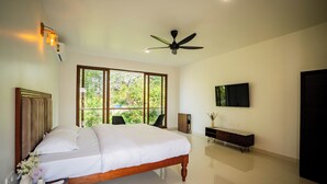 Un dormitorio con una cama, ventilador de techo, televisor montado en la pared y ventana con vista a la vegetación.