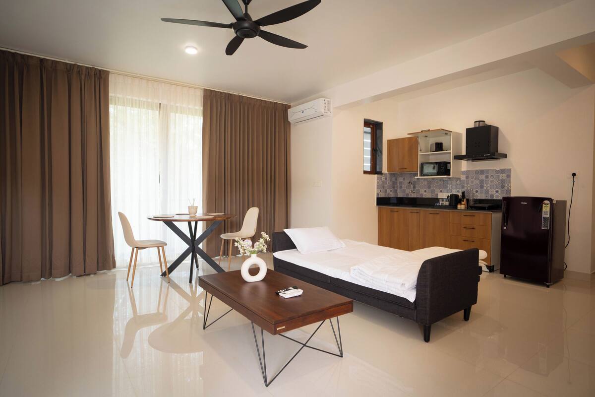 Una habitación de hotel moderna con cama, una mesa de comedor con sillas, una mesa de centro, un ventilador de techo, una zona de cocina con armarios y electrodomésticos, y un ventanal con cortinas.