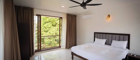 Un dormitorio amplio con una cama grande, un ventilador de techo, una ventana con cortinas y una mesita de noche con lámpara.