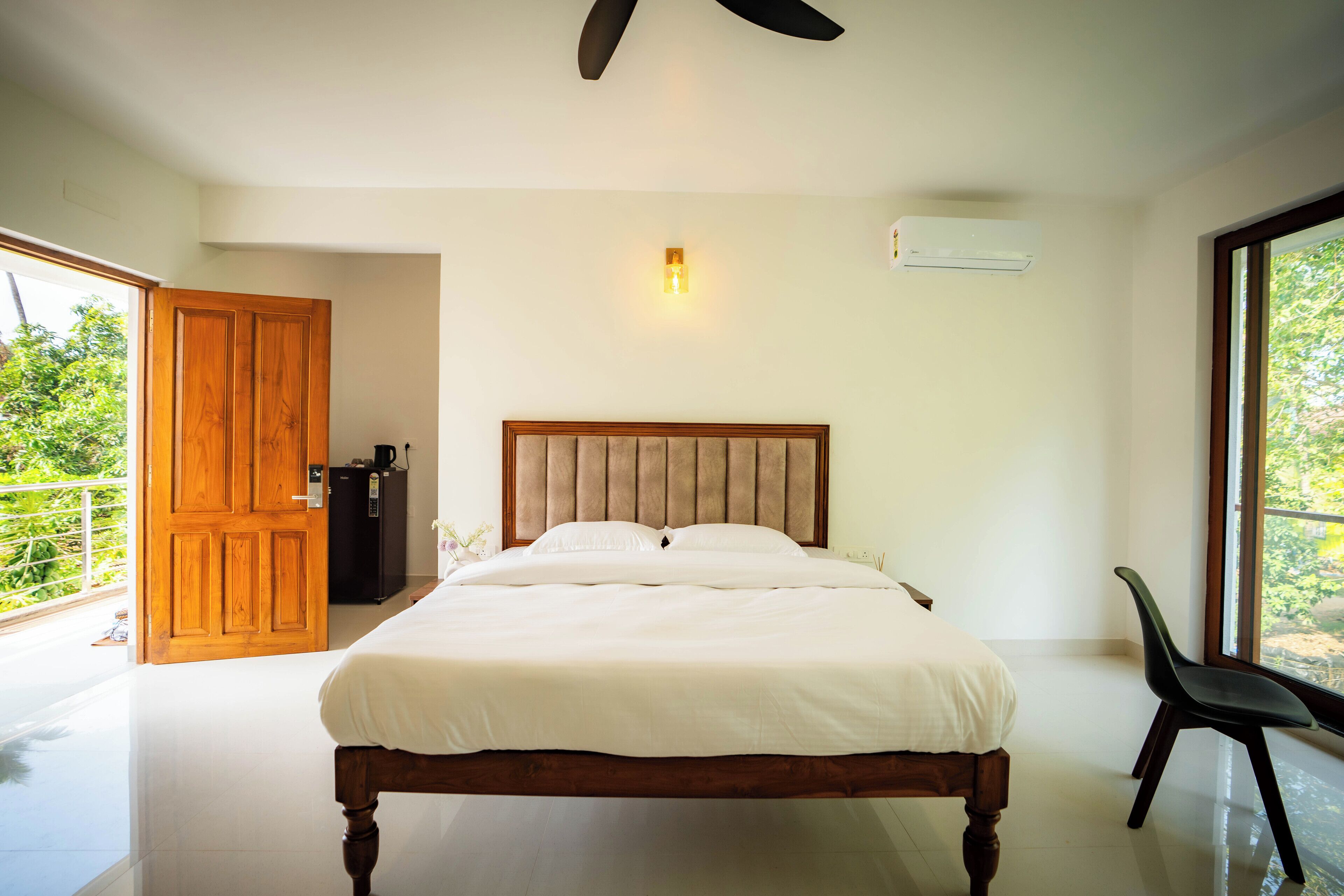 A bedroom with a wooden bed, a ceiling fan, a wall-mounted air conditioner, and a view of greenery through a large window.
