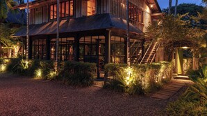 A large wooden house with multiple windows and a covered porch, surrounded by palm trees and a gravel path. - Rumah Hujan (Ubud)