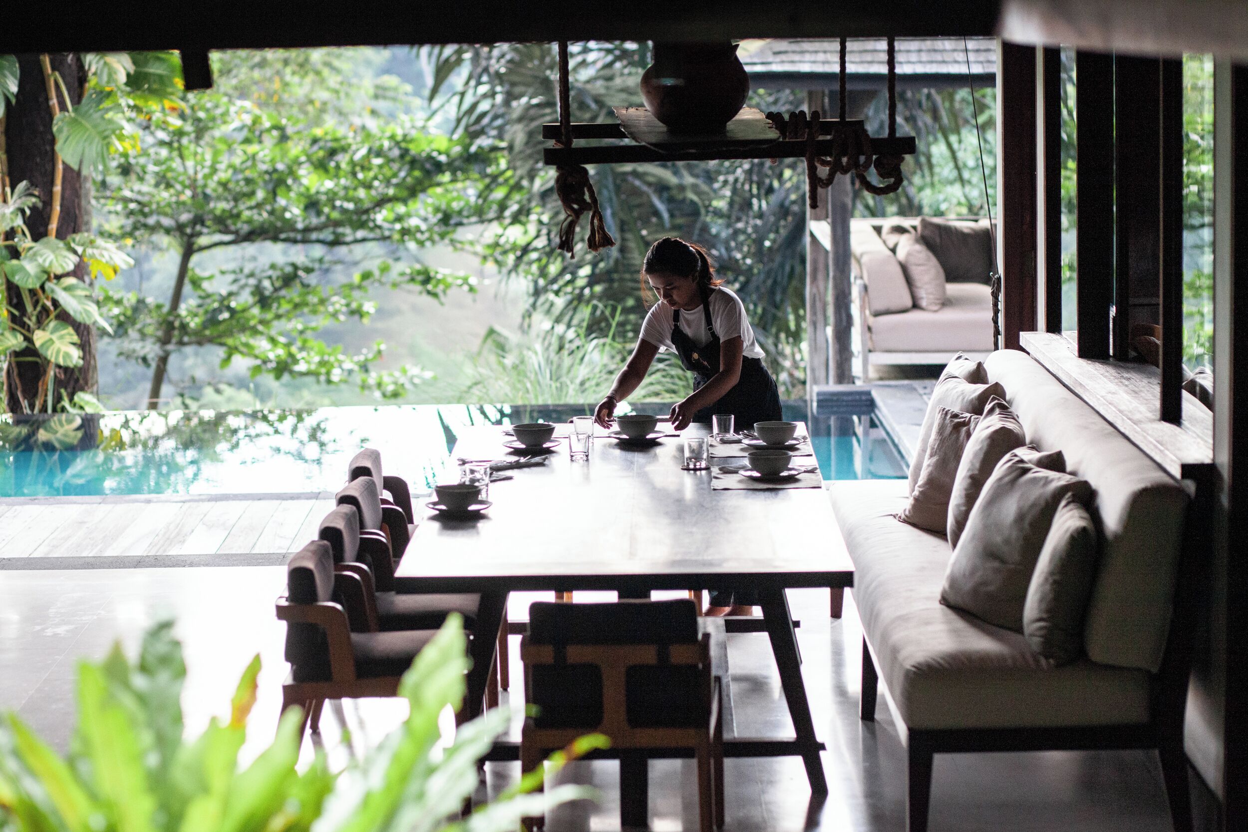 A dining area with a table set for a meal, a sofa, and a view of a pool and greenery.
