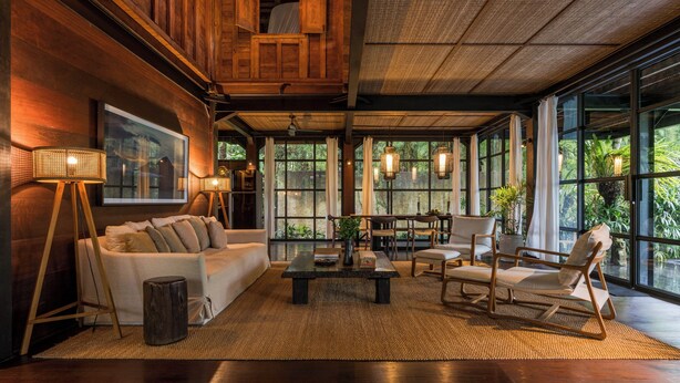 A spacious living area with a high ceiling, wooden paneling, a large window, and a comfortable seating arrangement. - Rumah Hujan (Ubud)