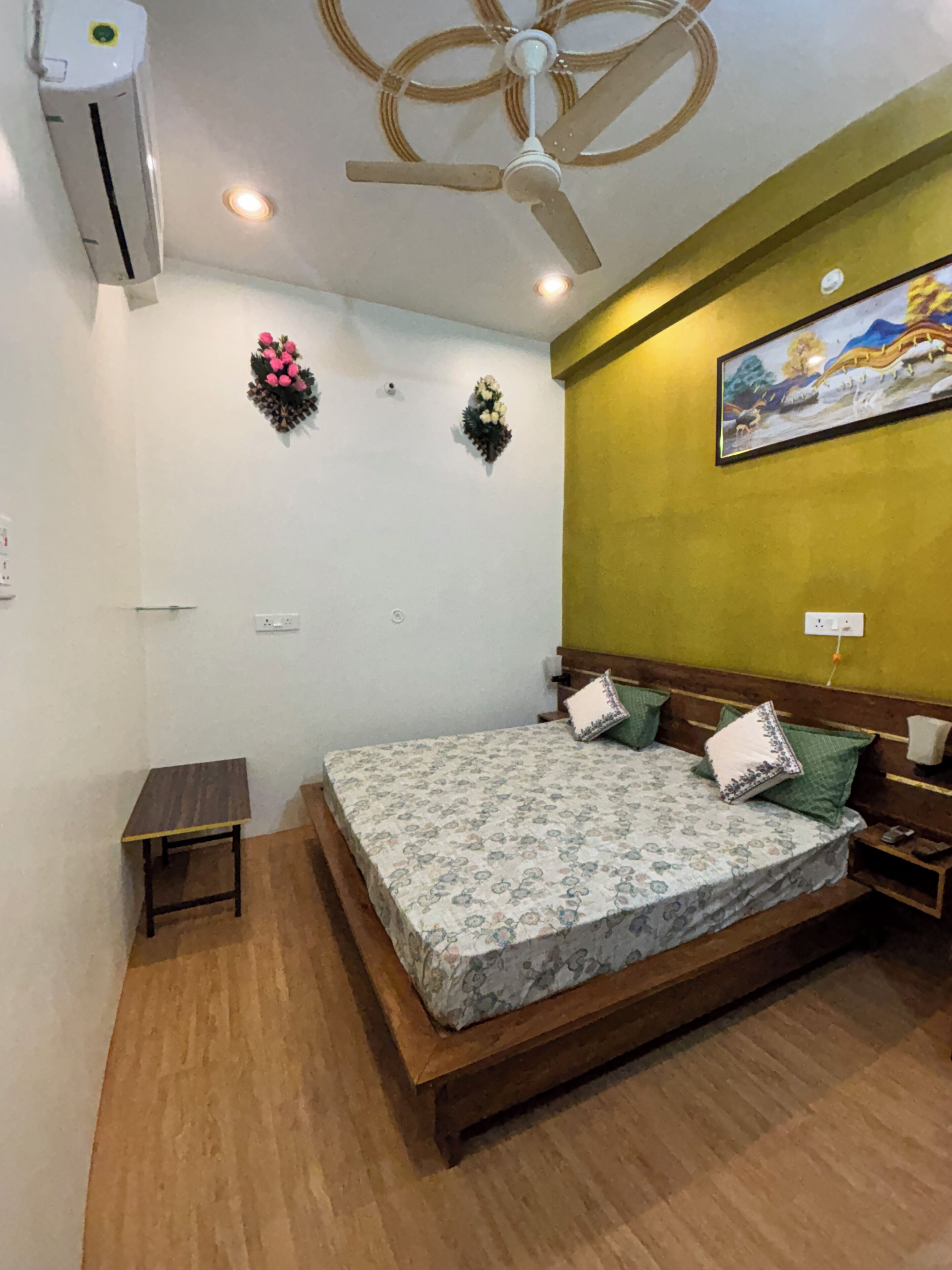 Un dormitorio con cama, una mesita de noche, una pequeña mesa, aire acondicionado, ventilador de techo y un cuadro colgado en la pared.