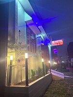 A modern hotel exterior with a neon sign, a well-lit entrance, and a landscaped garden area.